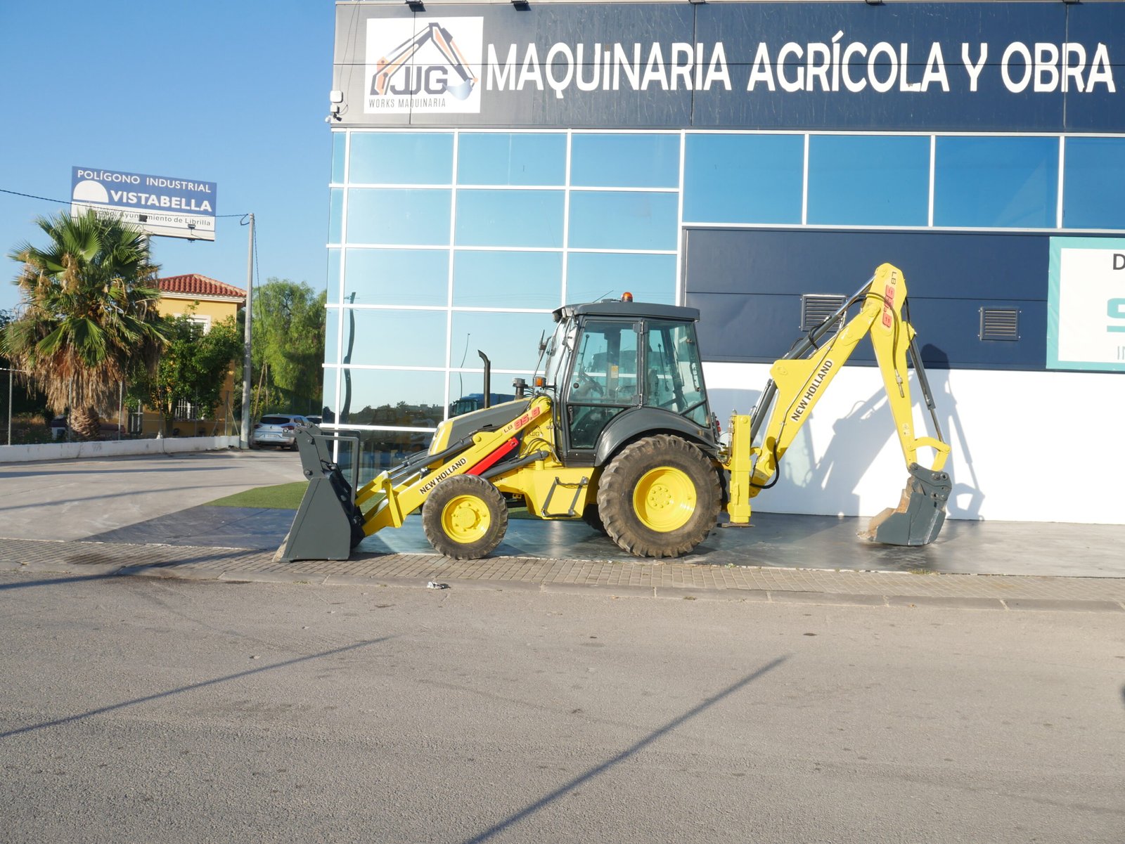 Retroexcavadora New Holland LB 95 B equipada con pinzas porta palets y aire acondicionado trabajando en entorno urbano